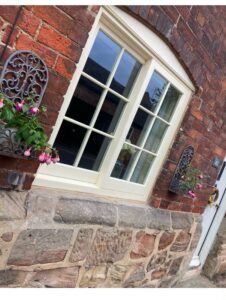 This image captures a beautifully preserved window with Georgian astragals, painted in off-white, contrasting against the rich red brickwork of an old building, complemented by decorative ironwork and blooming flowers.