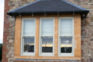 An architectural detail of a building featuring a triple sash window with wooden frames in sage green and white blinds, integrated seamlessly into a facade of rustic stone.