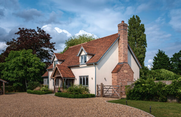 A charming new build home boasting an oak frame with striking red grandis timber windows, set against a verdant landscape and a pebbled driveway, under a dynamic cloudy sky.
