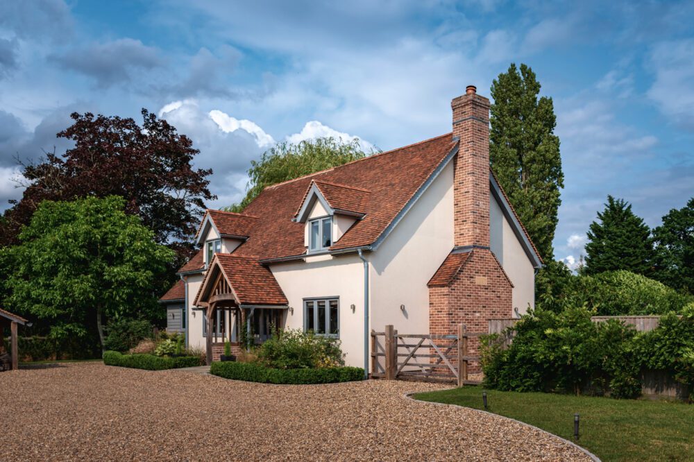 A charming new build home boasting an oak frame with striking red grandis timber windows, set against a verdant landscape and a pebbled driveway, under a dynamic cloudy sky.