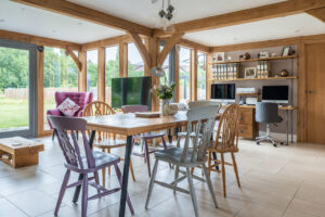 A welcoming dining space with a robust oak timber frame, bespoke red grandis timber windows, and a mix of classic and contemporary chairs around a wooden table.