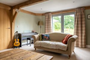 A peaceful music corner for relaxation, showcasing an upright piano, comfy seating, oak beams, and bespoke red grandis timber windows inviting in the garden views.