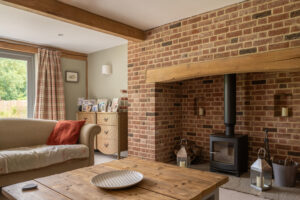 A cosy living room anchored by a traditional brick fireplace, complete with oak beams, bespoke red grandis timber windows, and a wooden coffee table.