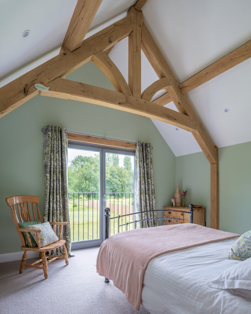 An airy and rustic bedroom featuring an elegant exposed oak timber frame, sage green walls, a rocking chair by the Juliet balcony window, and a view of the greenery outside.